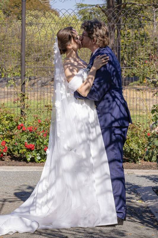 Bride and groom kissing in dappled garden light beside an iron fence and roses